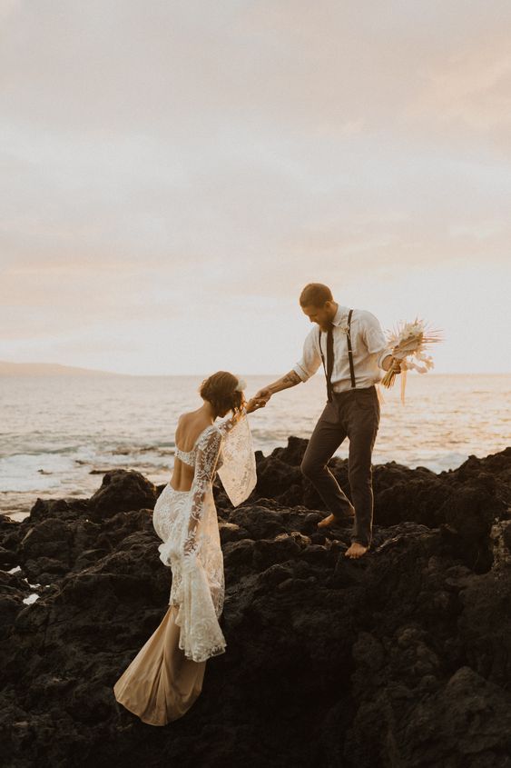 A bride and groom near the ocean, climbing on rocks, her in a boho wedding gown, him reaching back for her hand.