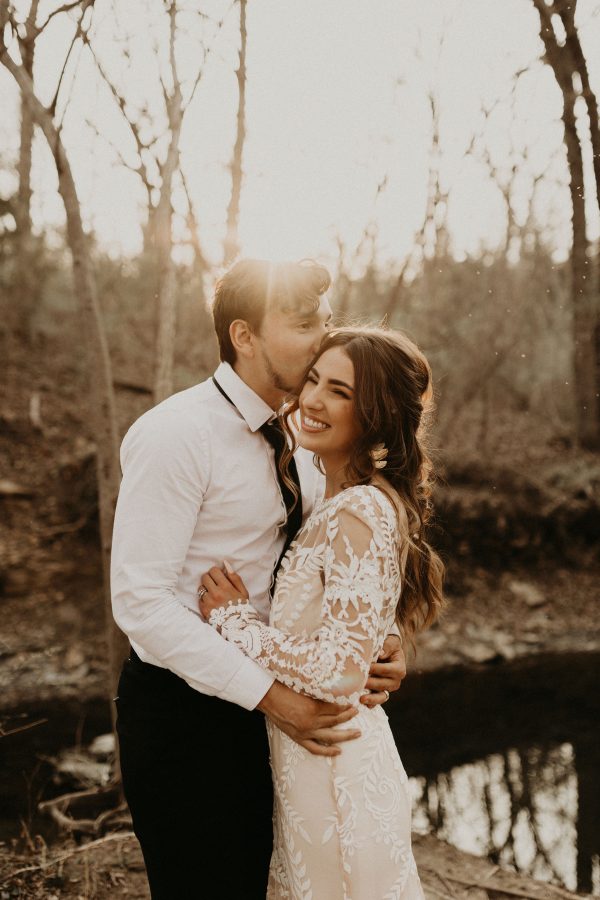 A groom kissing his bride on the forehead in the woods, bride in a boho lace wedding dress