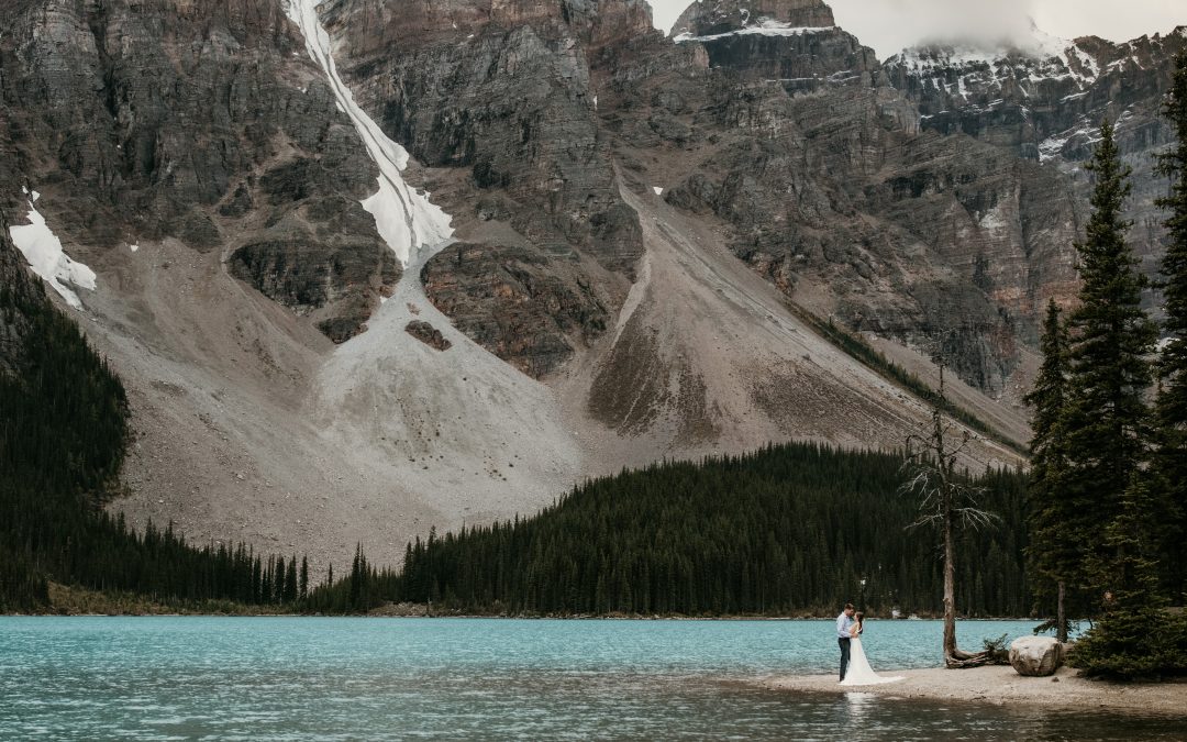 A bride and groom embracing under a glacier at Moraine Lake in Banff National Park