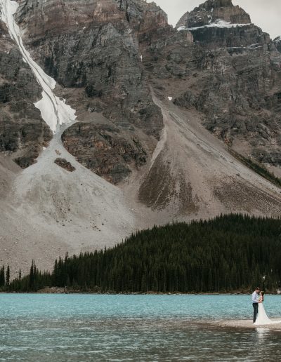 A bride and groom embracing under a glacier at Moraine Lake in Banff National Park