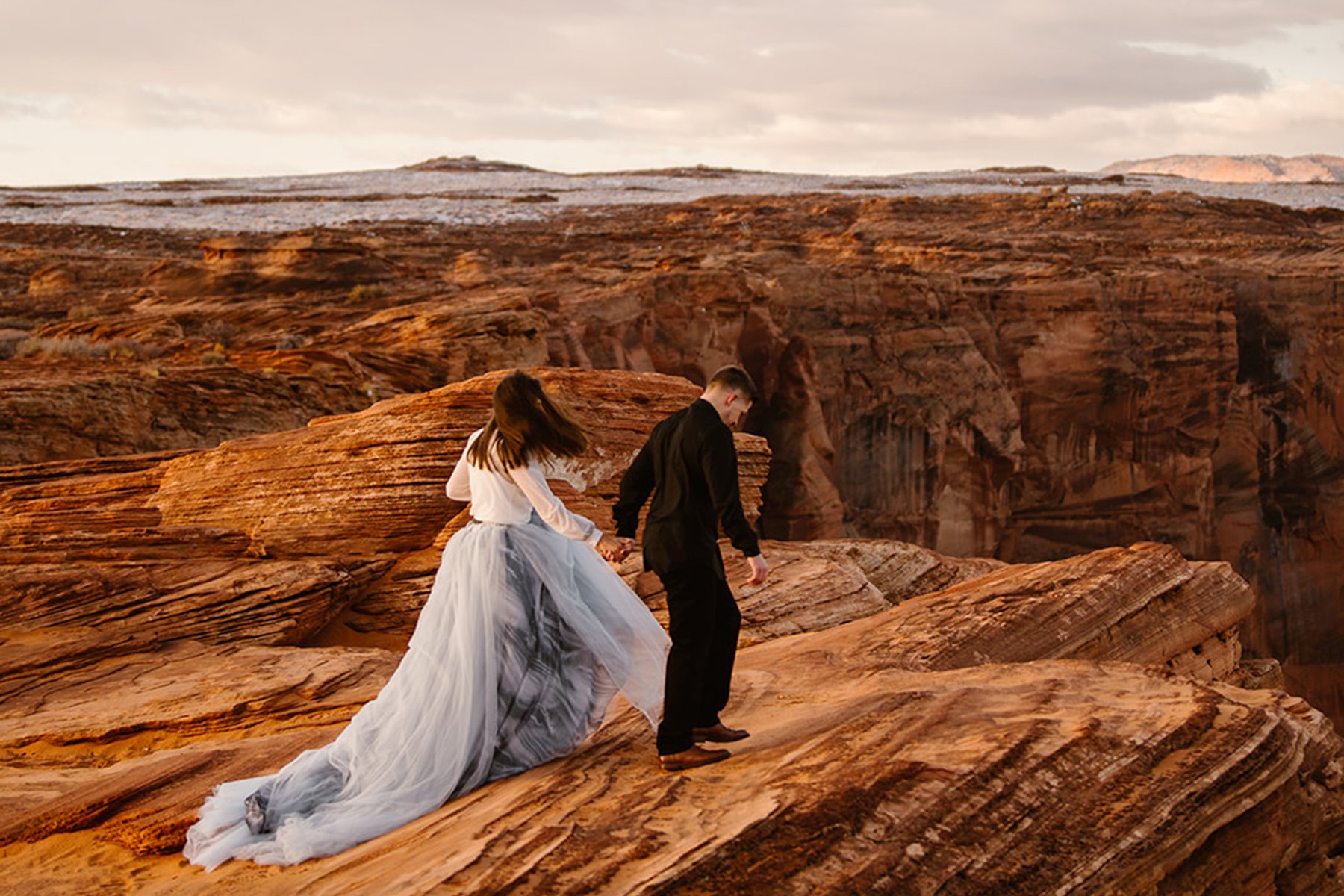 A couple walking on the orange cliffs in Arizona holding hands. Woman is wearing a dramatic blue painted tulle skirt.