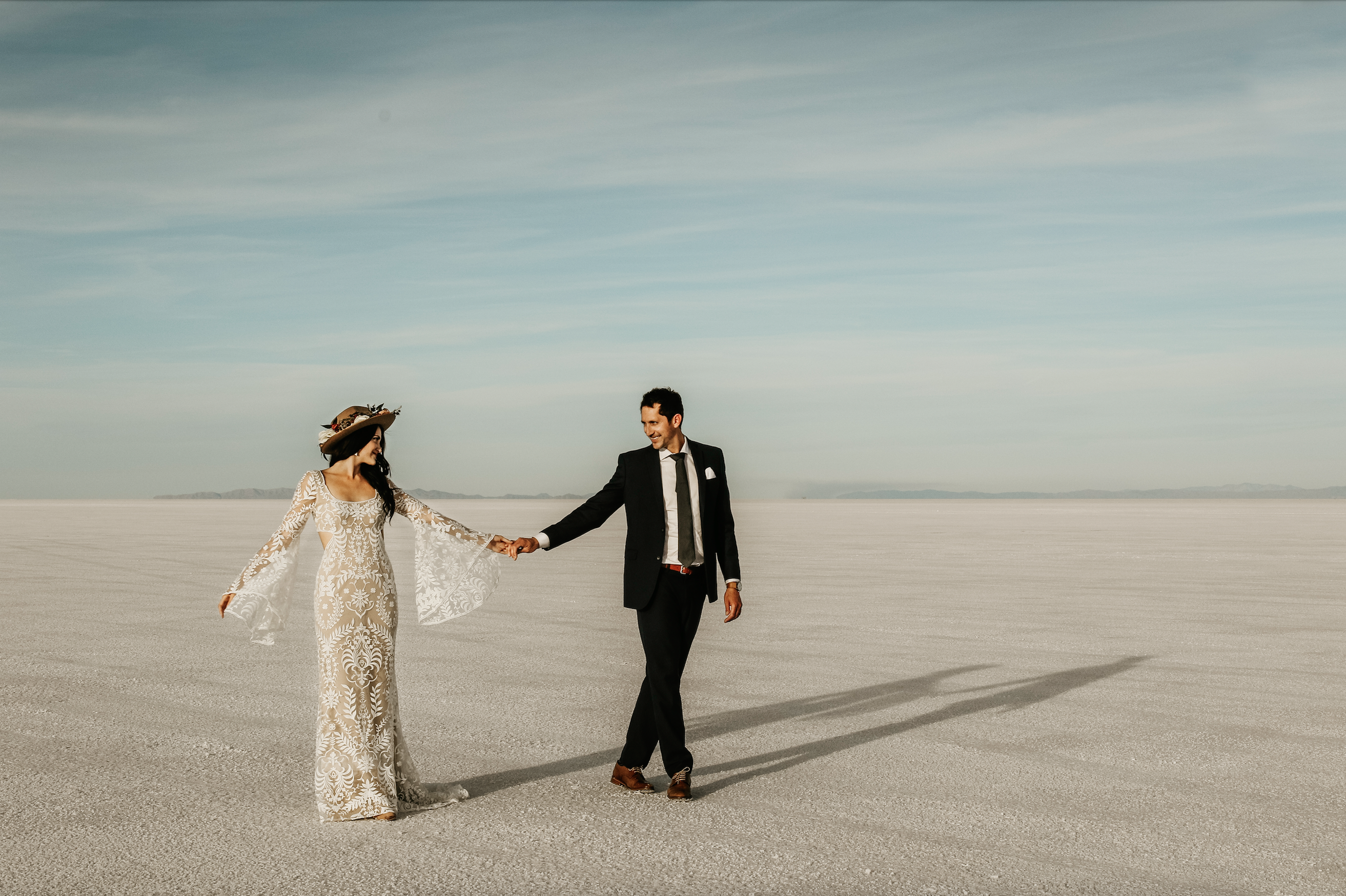 A bride and groom holding hands on the salt flats in Utah, her wearing a designer wedding gown and hat, him in a suit.