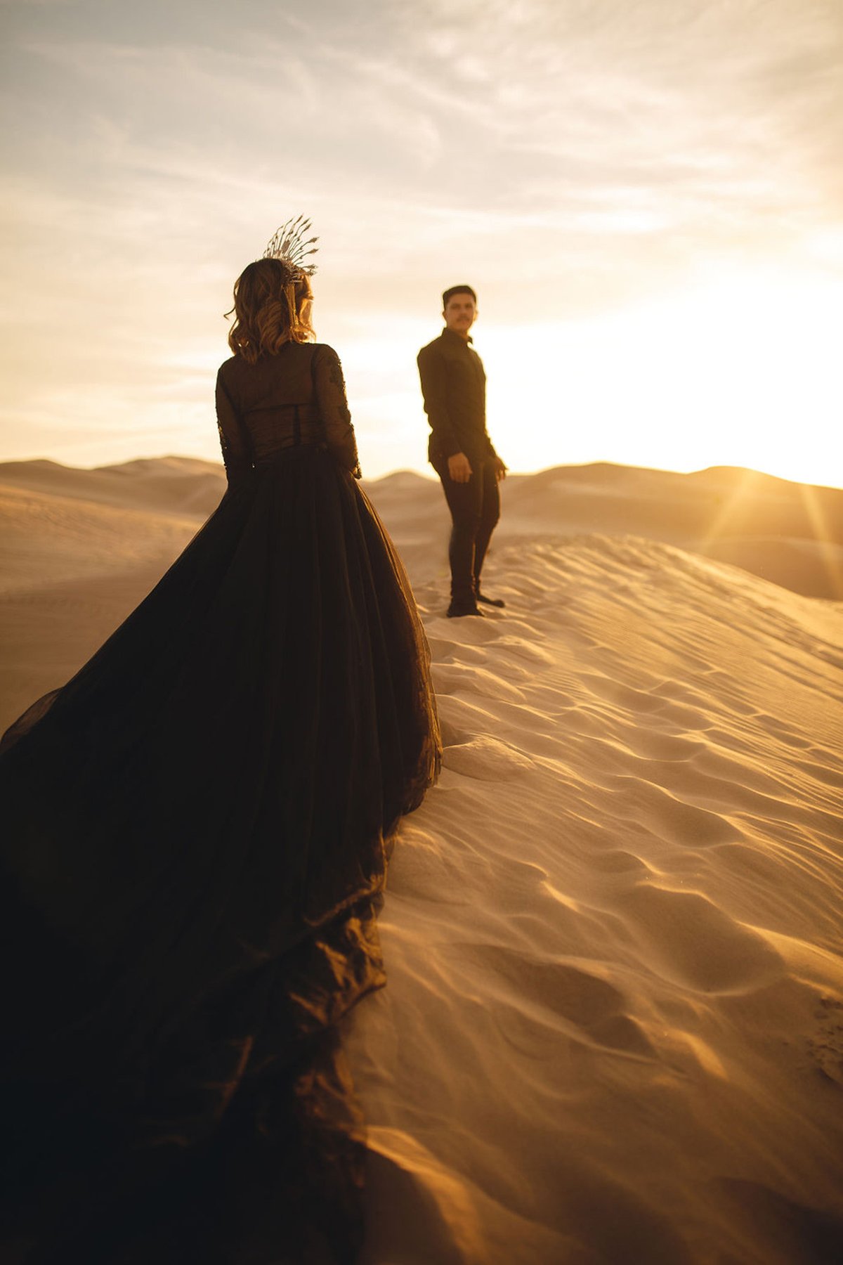 A couple dressed in black, standing in the dunes at sunset, he is looking back at her wearing a long train black tulle dress and crown