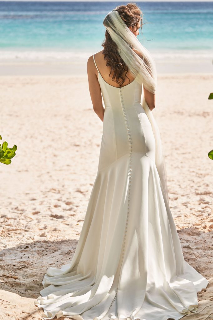 A gorgeous bride on a beach wearing a simple spagetti strap wedding gown with buttons all the way down the train