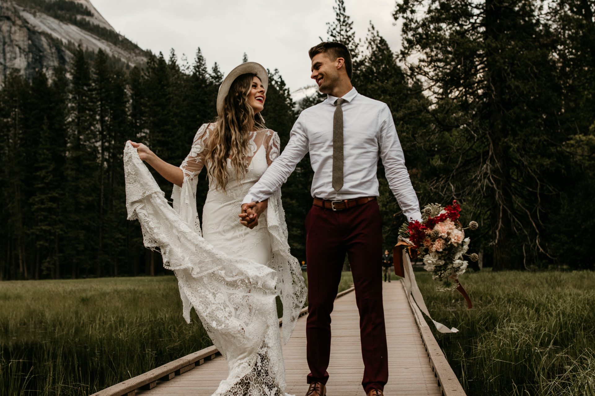 A bride and groom, her in a designer wedding gown by Rue de Seine, walking across a wooden bridge in Yosemite National Park for a styled photo shoot.