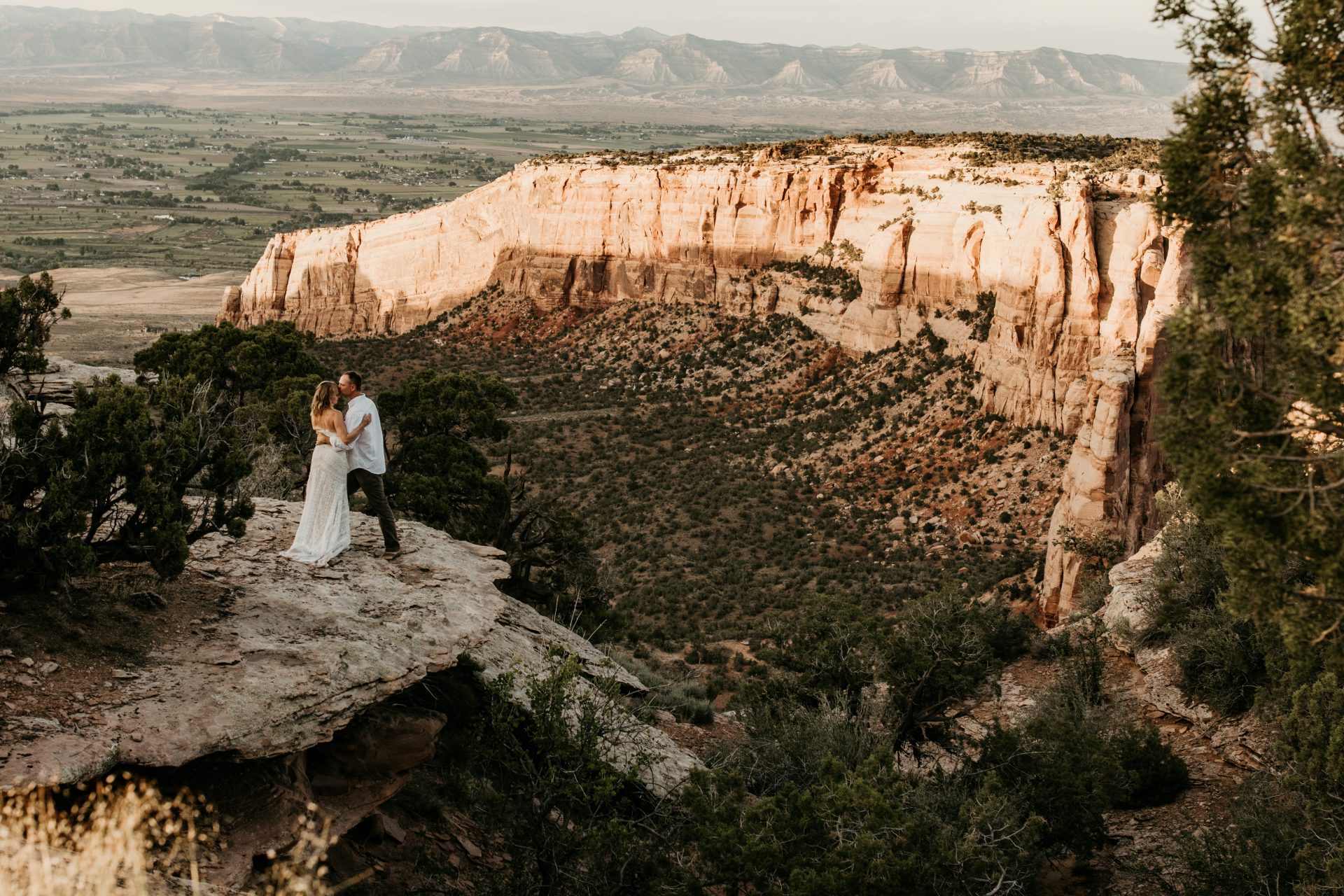 A bride and groom standing on the edge of a cliff kissing at sunset with the mountainous landscape behind them