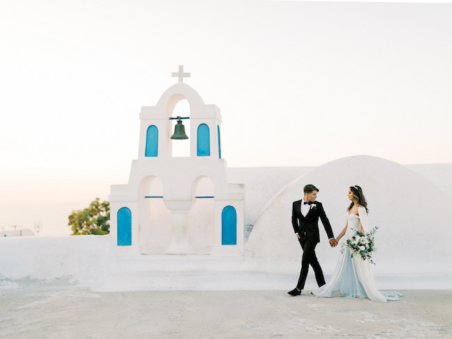 Image of a bride and groom eloping in front of a whitewashed chapel in Greece. Mixing religion and beliefs with romance and a sacred adventure.